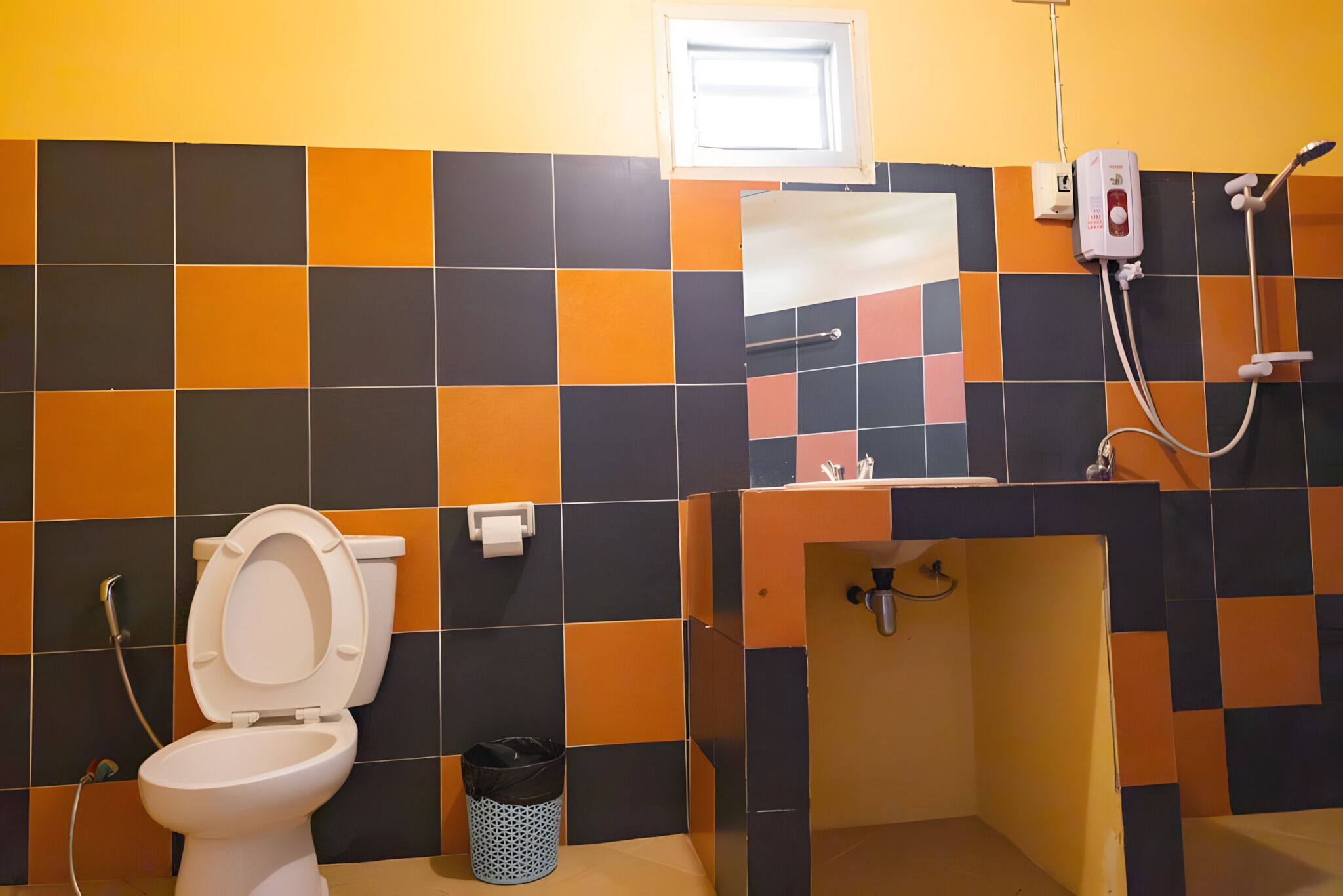 A bathroom with a toilet, a trash bin, a shower, and a tiled wall with a window.