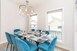 A dining area with a glass table set for six, blue upholstered chairs, and a modern chandelier. - Where Luxury Feels Like Home | Cozy, Comfy and Spacious 4-Bedroom Retreat (Edmonton)