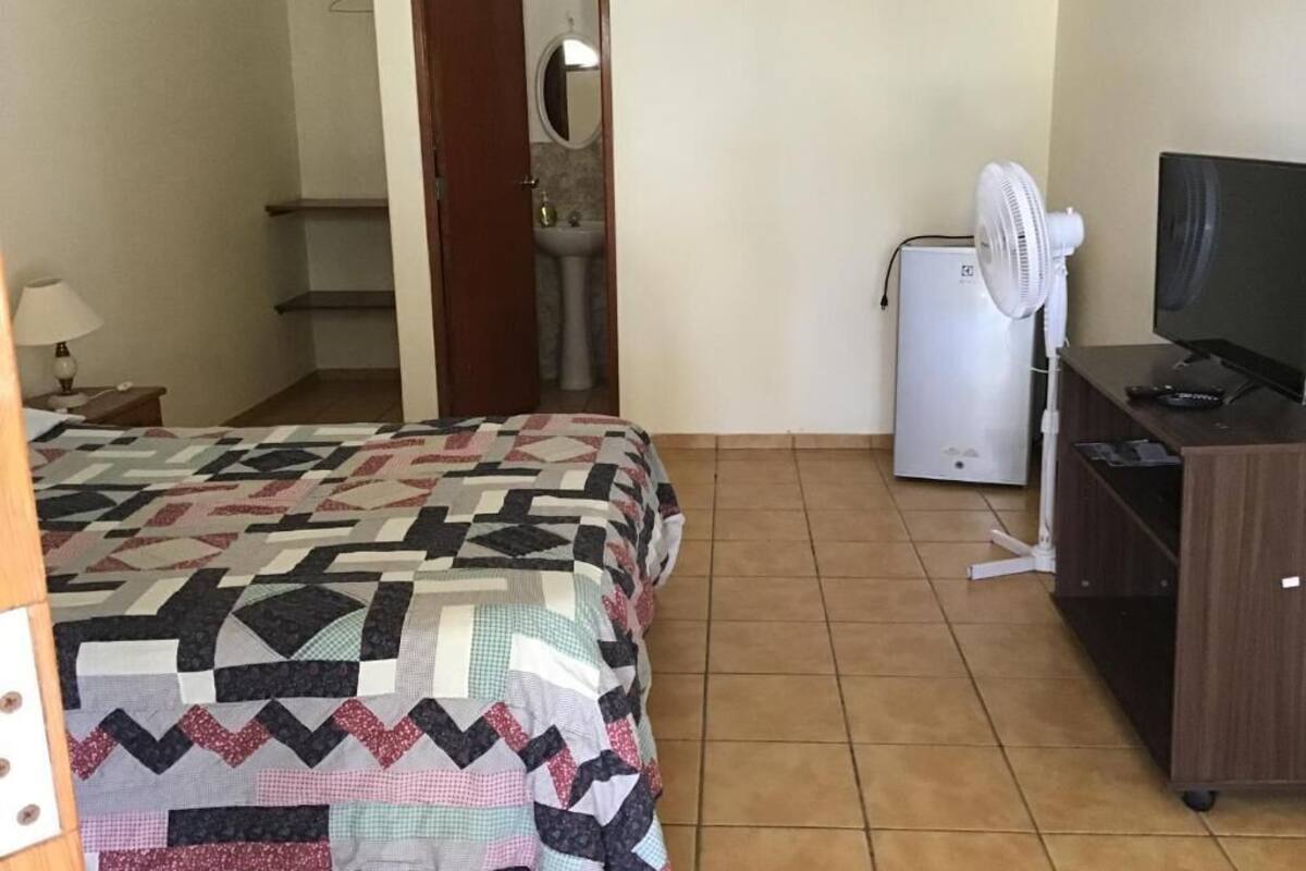 A bedroom with a bed, a desk with a computer, a fan, and a TV.