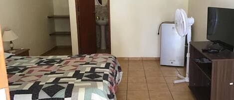 A bedroom with a bed, a desk with a computer, a fan, and a TV.