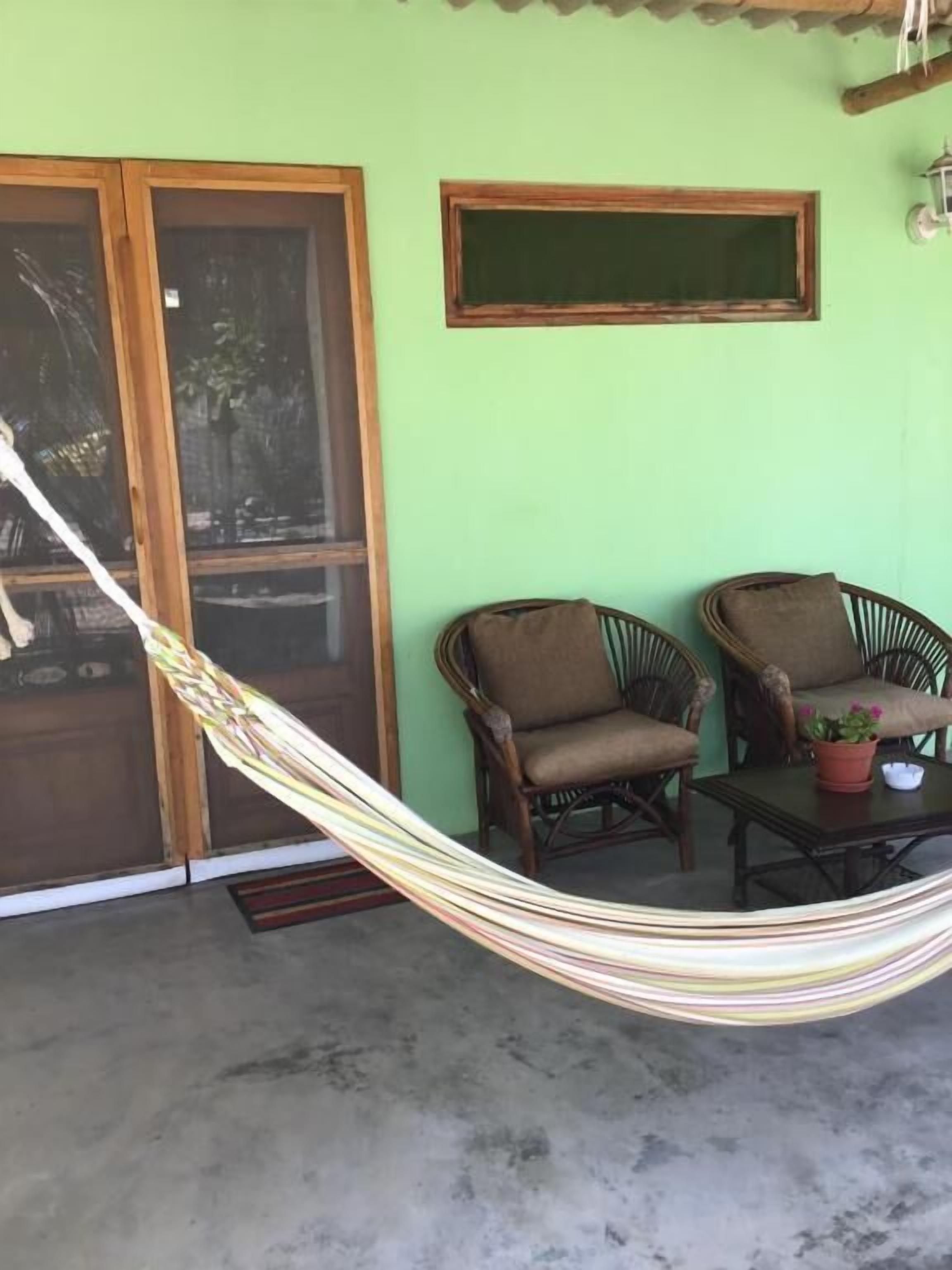 A room with a hammock, two wicker chairs, a small table with a potted plant, a window, and a door.
