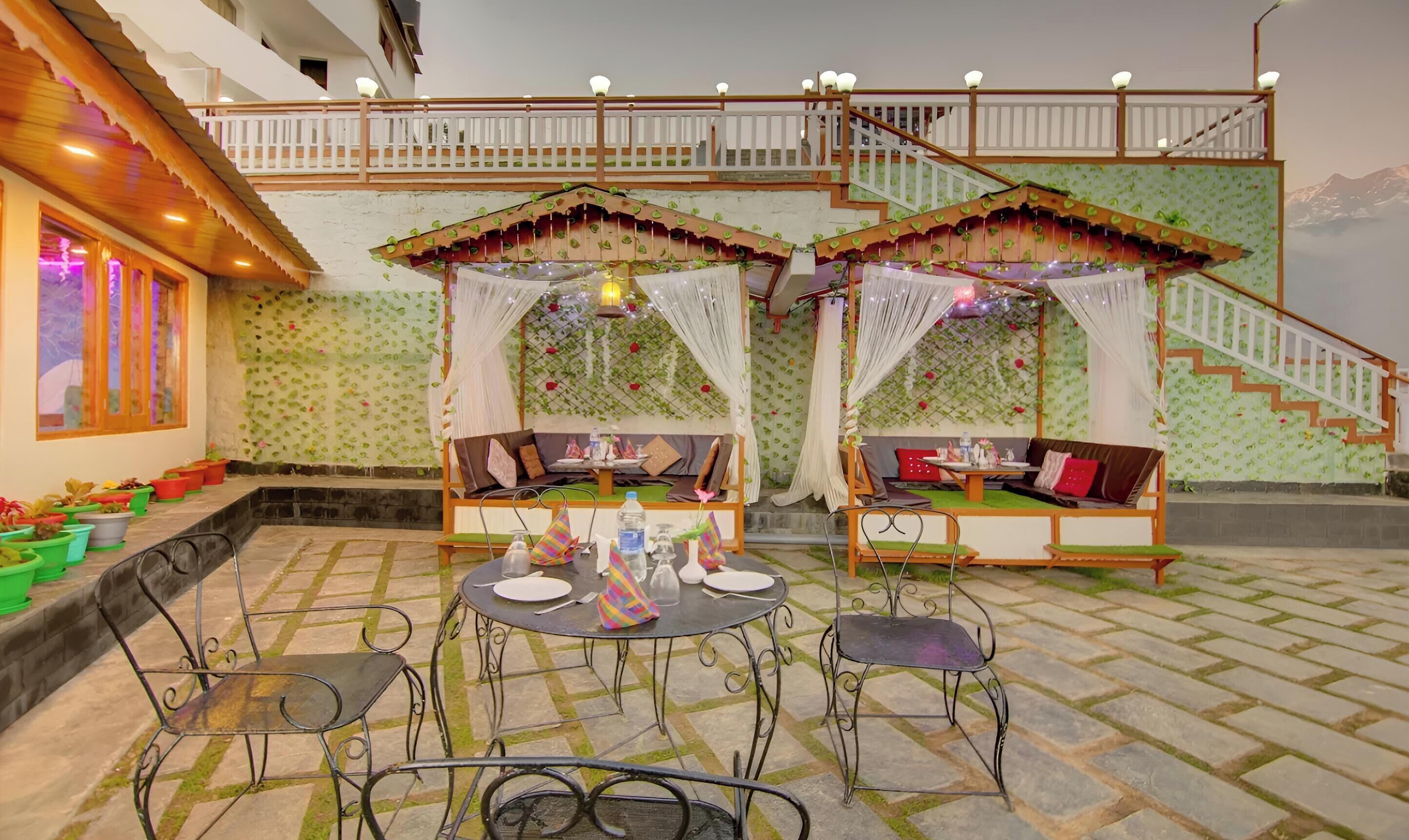 A terrace with two wooden benches, a table, and chairs, surrounded by potted plants.