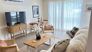 A modern living room with a flat-screen TV, wooden furniture, a dining table, and a large window with sheer curtains. - Coral Beach #204 - by ZenBreak (Little Battaleys)