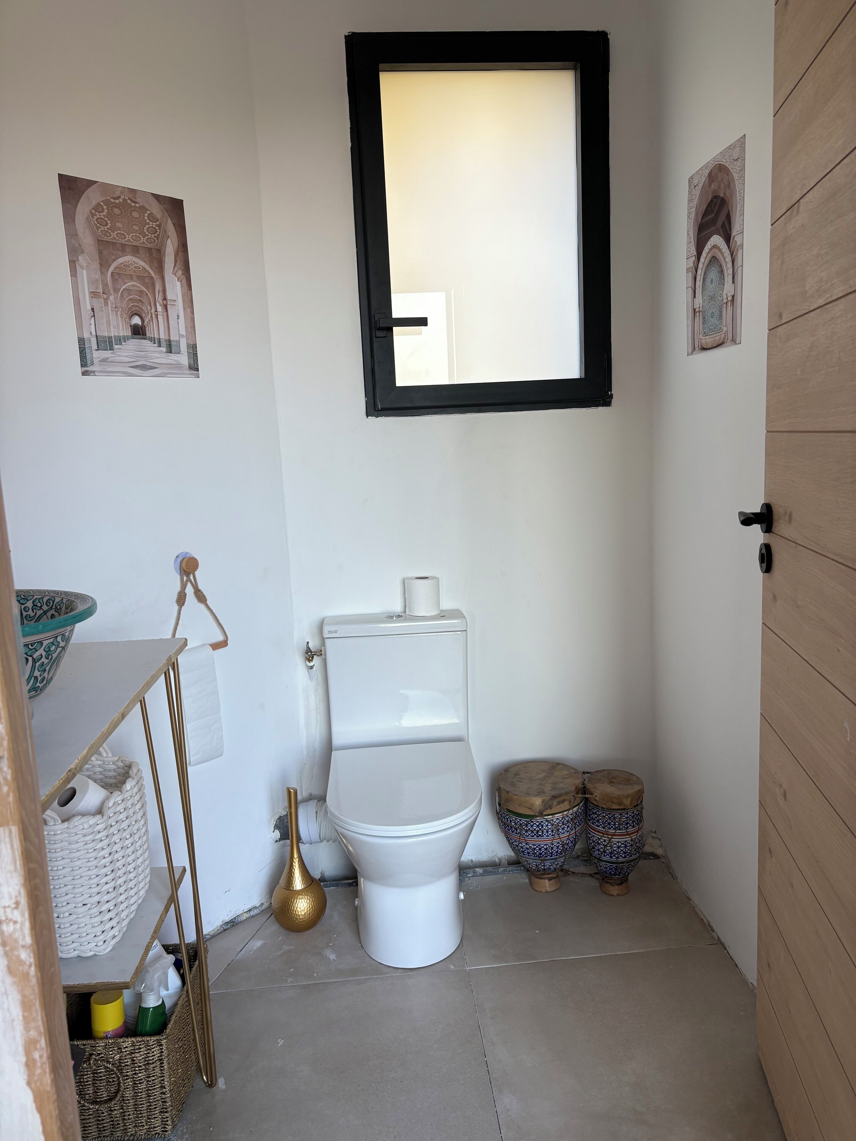 A small bathroom with a toilet, a window, and a shelf with a bowl and a basket.