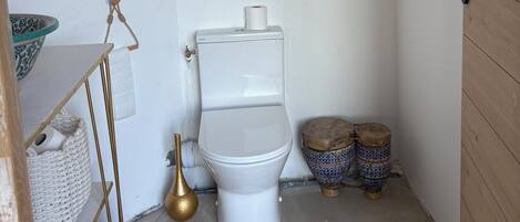 A small bathroom with a toilet, a window, and a shelf with a bowl and a basket.