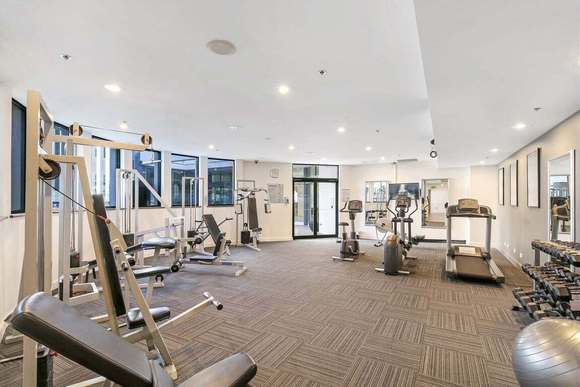 Un gimnasio moderno con diversos equipos de ejercicio, ventanas amplias y suelo alfombrado.