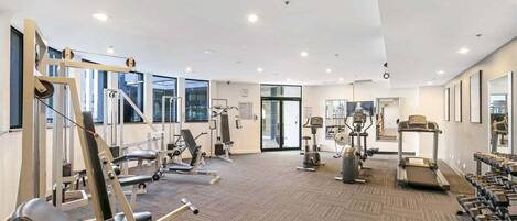 A modern gym with various exercise equipment, large windows, and a carpeted floor.