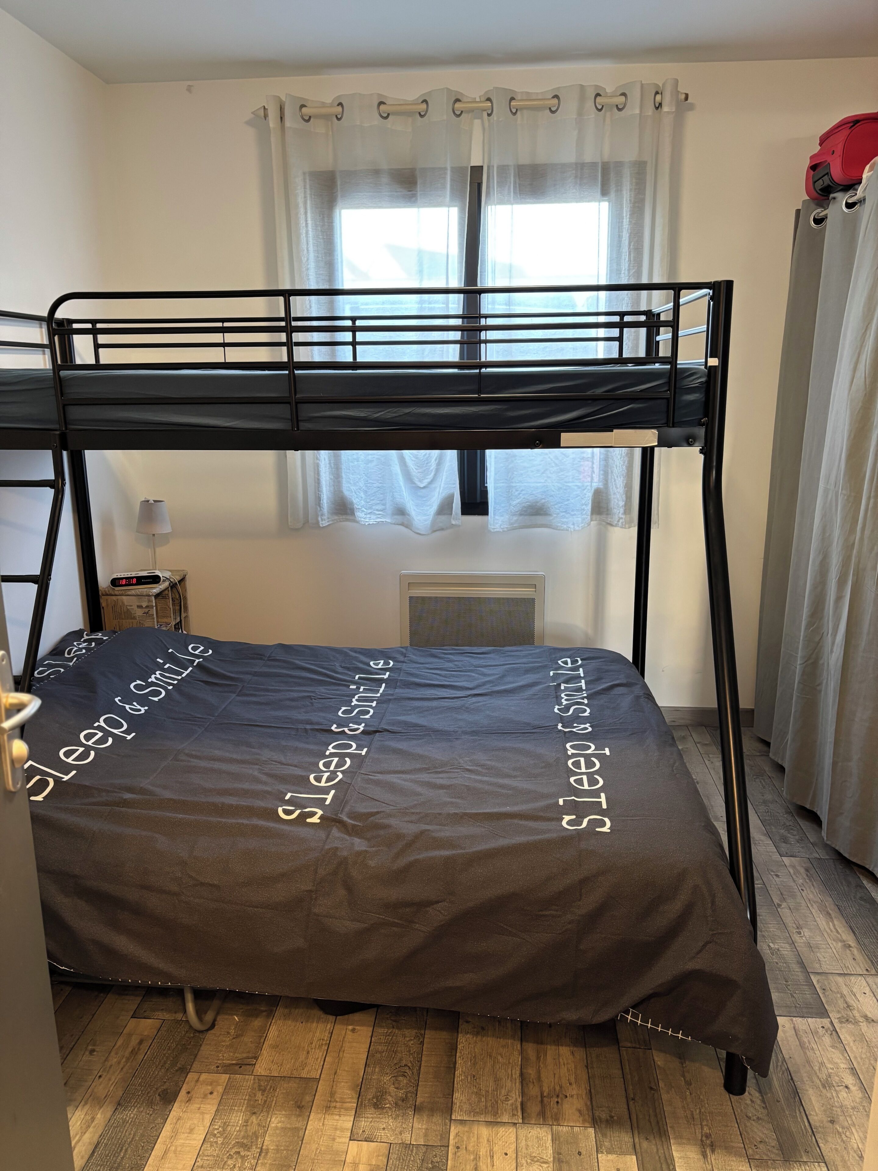 Un lit superposé avec un matelas sur lequel est imprimé le texte « dormez et souriez », une fenêtre avec des rideaux, un radiateur et un sac rouge posé sur le cadre du lit.
