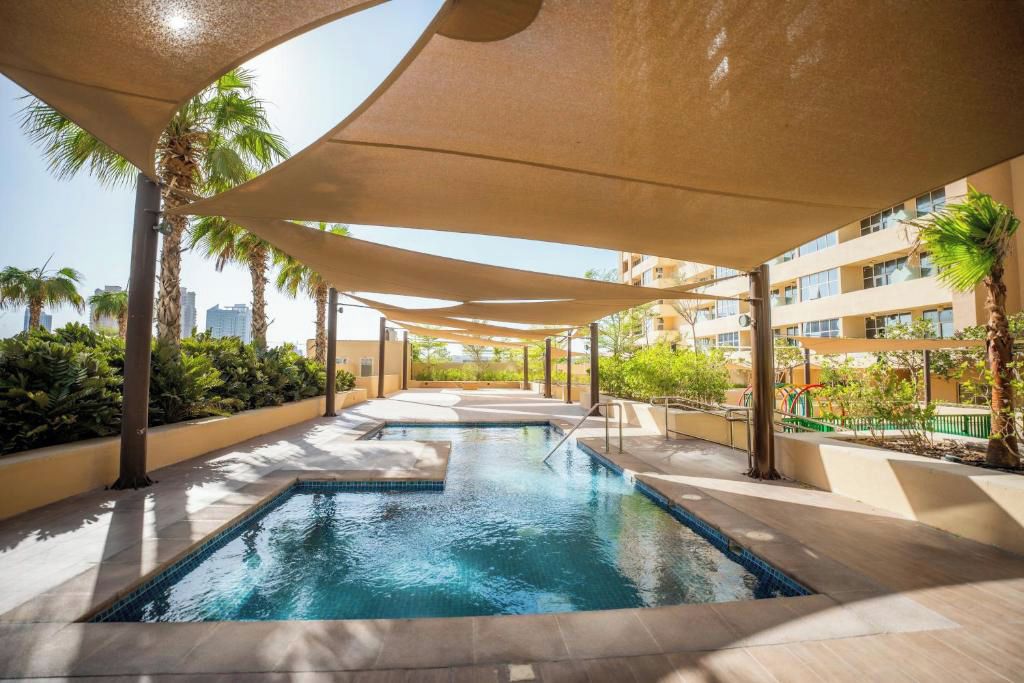 A swimming pool with a sunshade structure, surrounded by greenery and buildings.