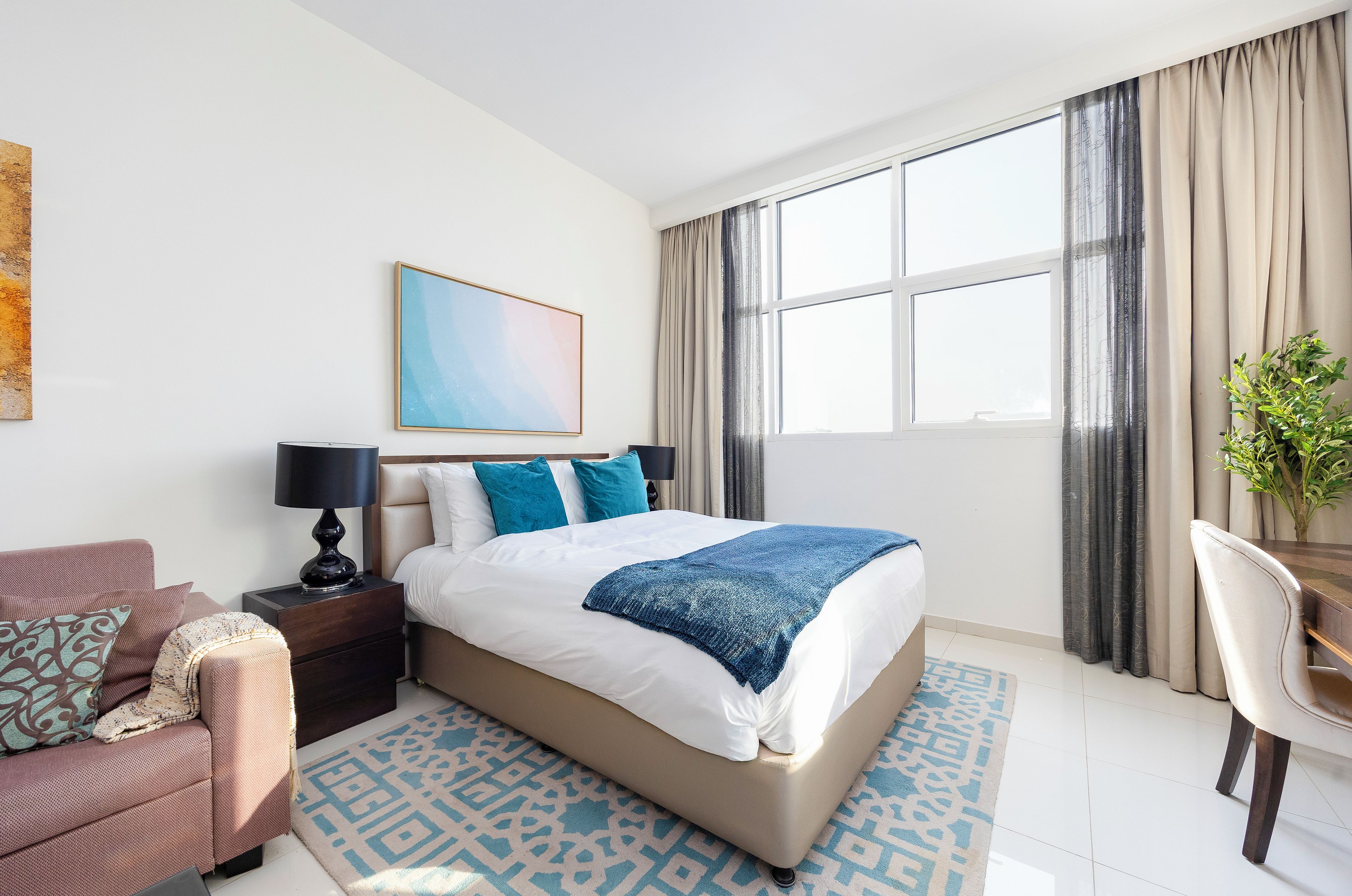 Habitación de hotel con cama, escritorio, silla, mesita de noche, lámpara, un cuadro y una ventana con cortinas.