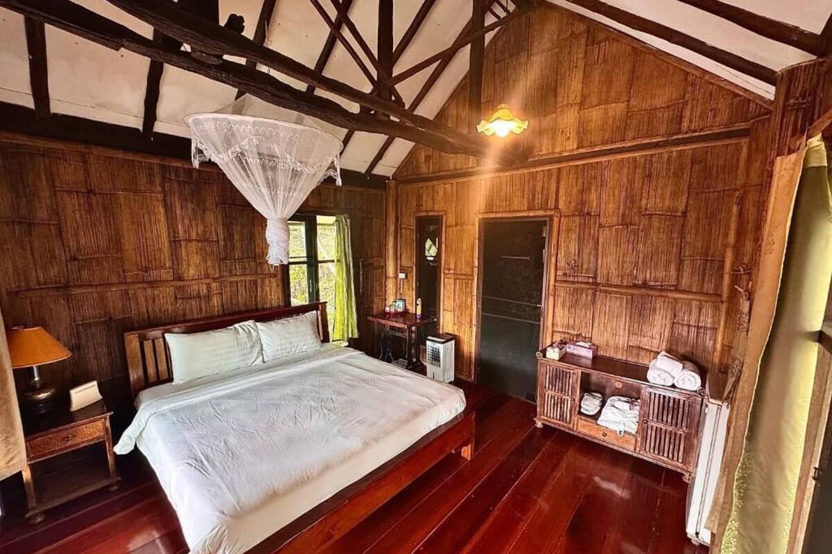 A wooden room with a bed, a nightstand, a bedside table, a lamp, a hanging light fixture, and a small cabinet with towels.
