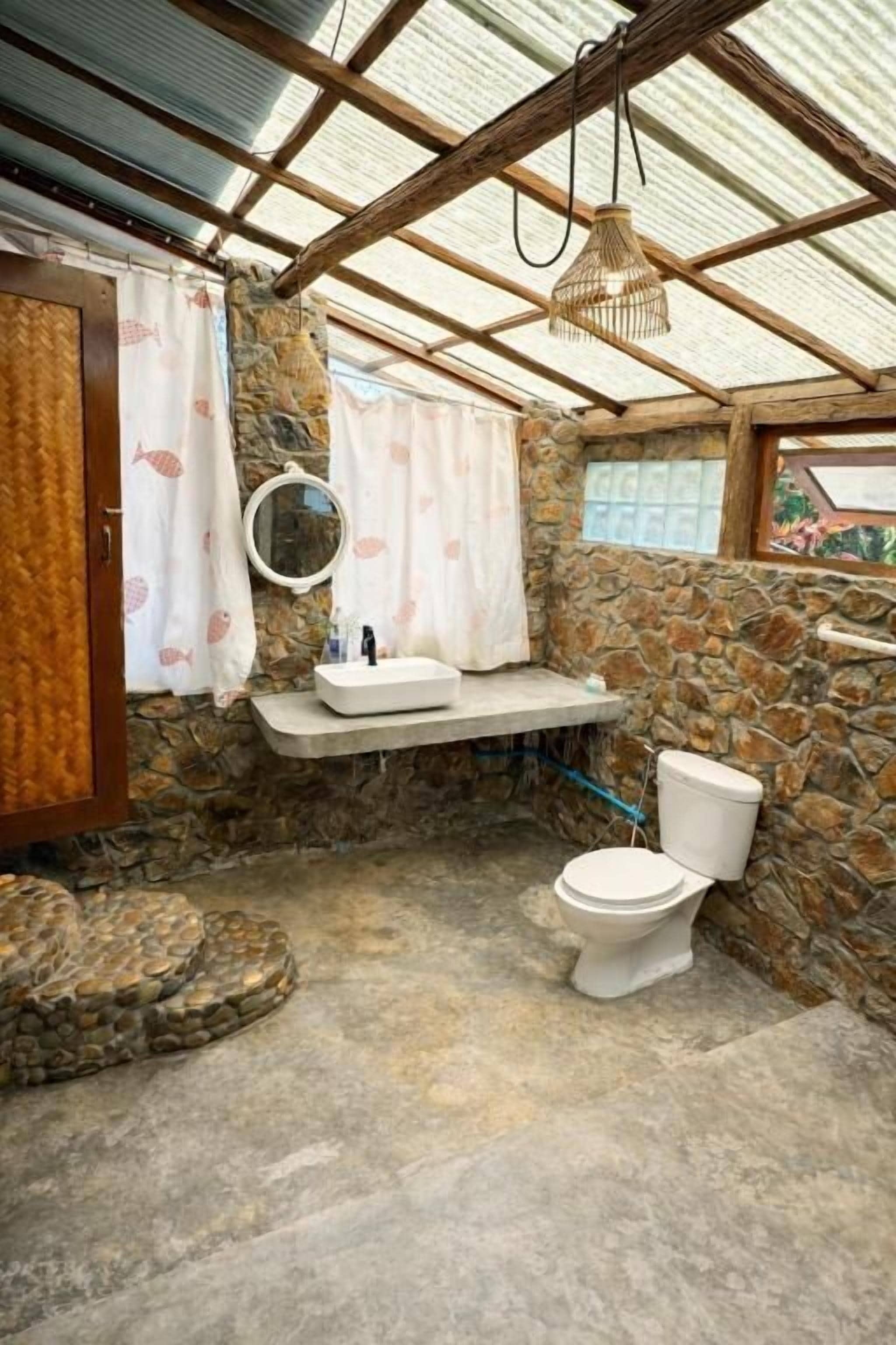 A bathroom with a stone wall, a wooden ceiling, a white sink, a toilet, and a hanging light fixture.