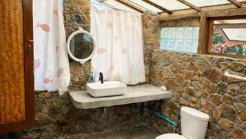 A bathroom with a stone wall, a wooden ceiling, a white sink, a toilet, and a hanging light fixture.