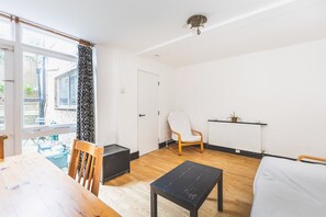 Ein Zimmer mit Holzboden, einem weißen Stuhl, einem schwarzen Tisch, einem Heizkörper und einem Fenster mit Vorhang.