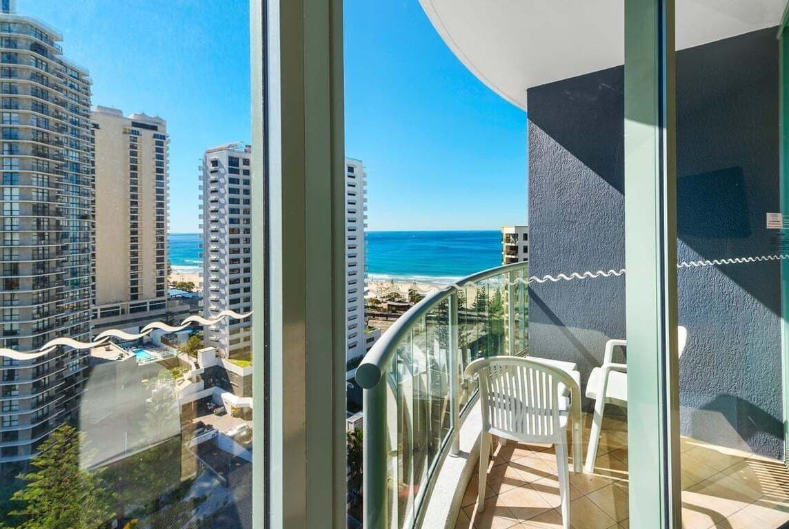 Un balcón con vista al océano y a la playa, barandilla de vidrio y una silla blanca.
