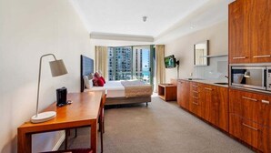 A hotel room with a bed, a desk, a TV, a kitchen area, and a large window with a city view. - Skyline Stay with Balcony (Gold Coast)