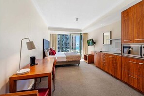 Ein Hotelzimmer mit Bett, Schreibtisch, Fernseher, Küchenzeile und großem Fenster mit Blick auf die Stadt.