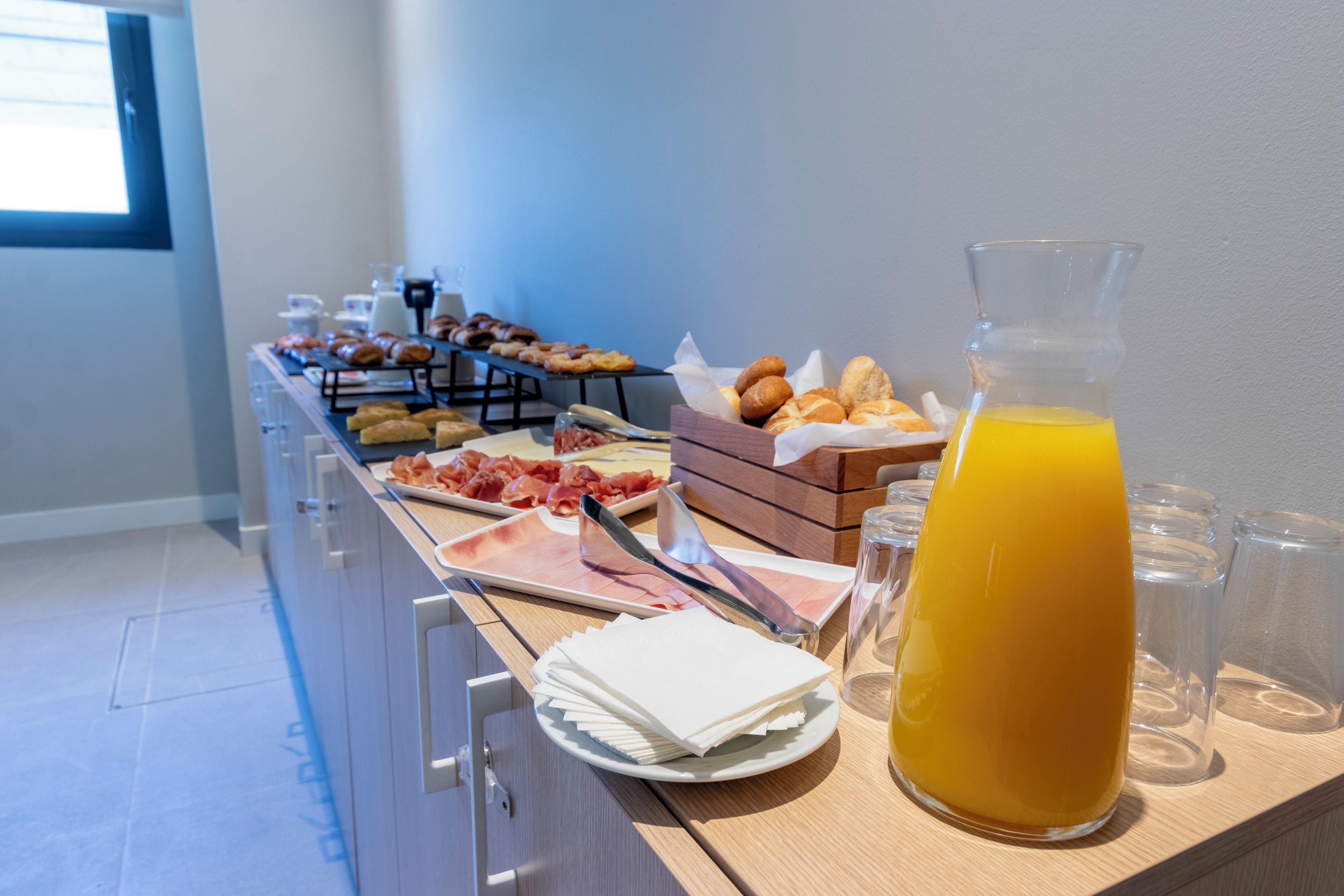 Un bufé con diversos alimentos, un dispensador de zumo y servilletas sobre una mesa de madera.