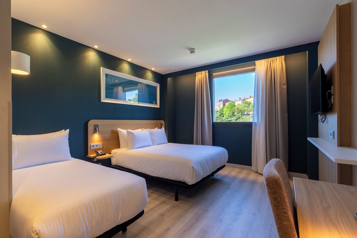 Quarto de hotel com duas camas, uma escrivaninha, uma cadeira, uma televisão, uma janela com cortinas e um aplique na parede.