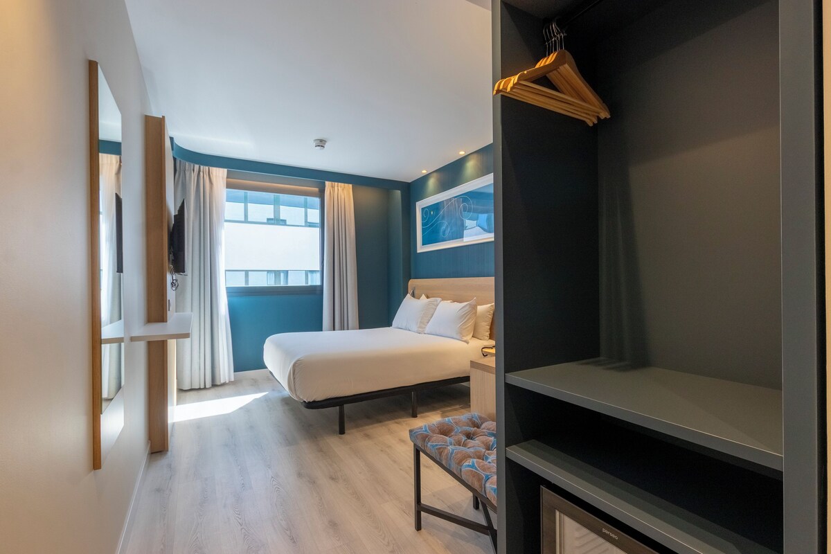 A modern hotel room with a bed, a bench, a TV, and a window with curtains.