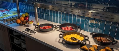 A buffet counter with various food items, including fruits, meats, and scrambled eggs, with a tiled backsplash and a view of a pool.