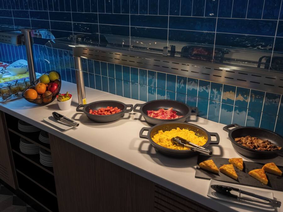 Mostrador de buffet con diversos alimentos, como frutas, carnes y huevos revueltos, un respaldo de azulejos y vista a una piscina.