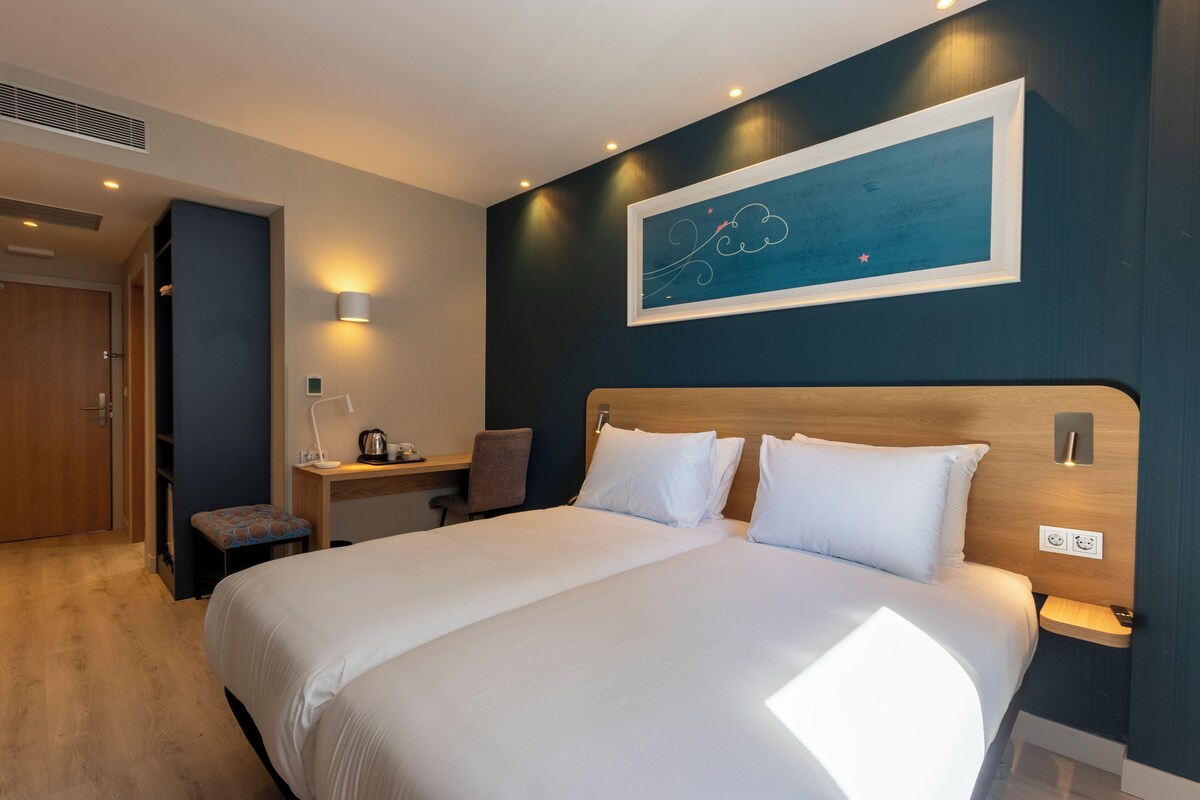 Une chambre d’hôtel avec un grand lit, un bureau avec une chaise, une œuvre d’art accrochée au mur et un petit banc.