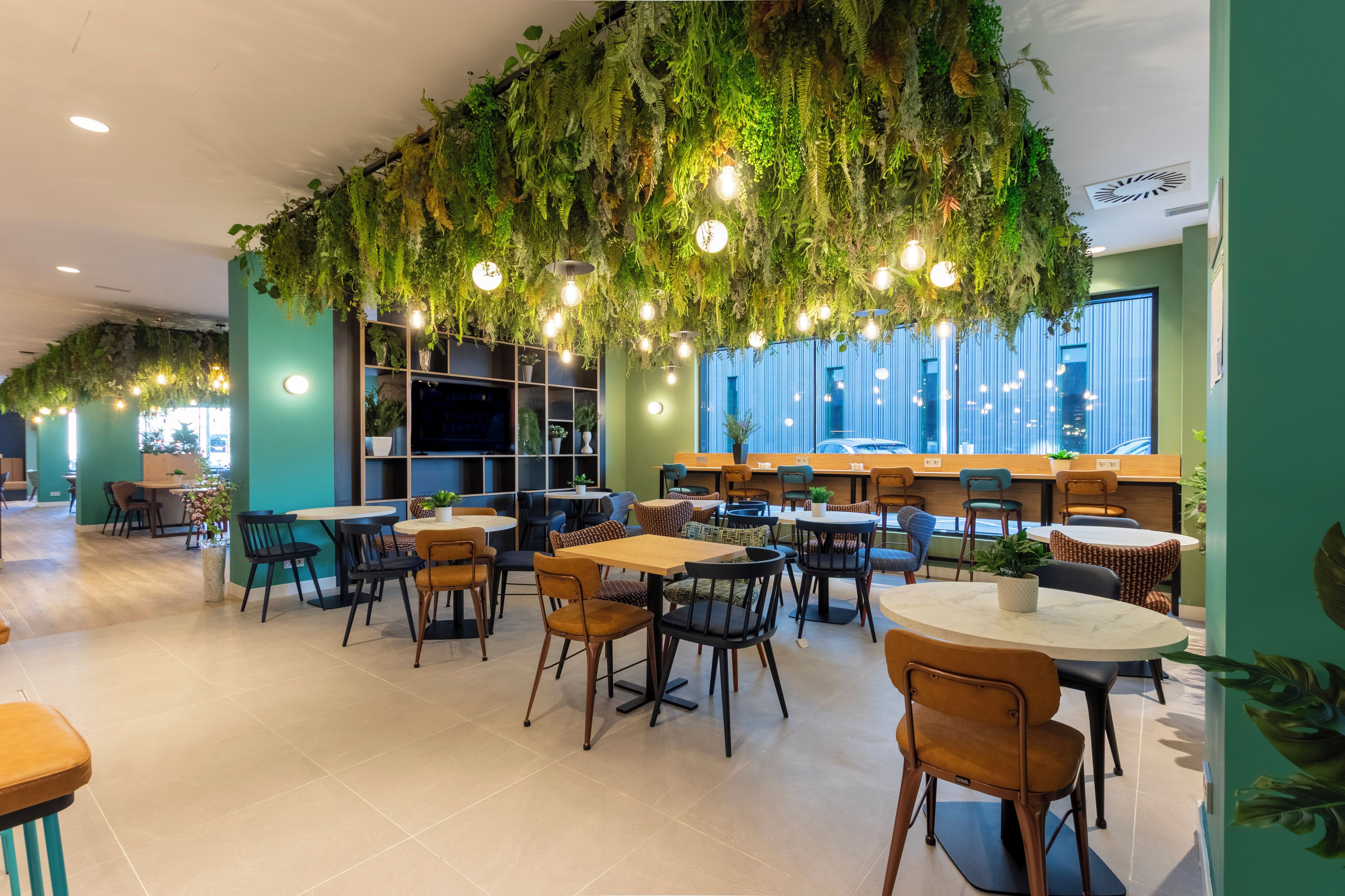 Un área de comedor interior moderna con mesas y sillas, una zona de bar, y un techo decorado con plantas y luces colgantes.