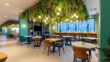 Un área de comedor interior moderna con mesas y sillas, una zona de bar, y un techo decorado con plantas y luces colgantes.
