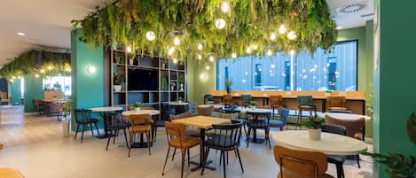 Un área de comedor interior moderna con mesas y sillas, una zona de bar, y un techo decorado con plantas y luces colgantes.