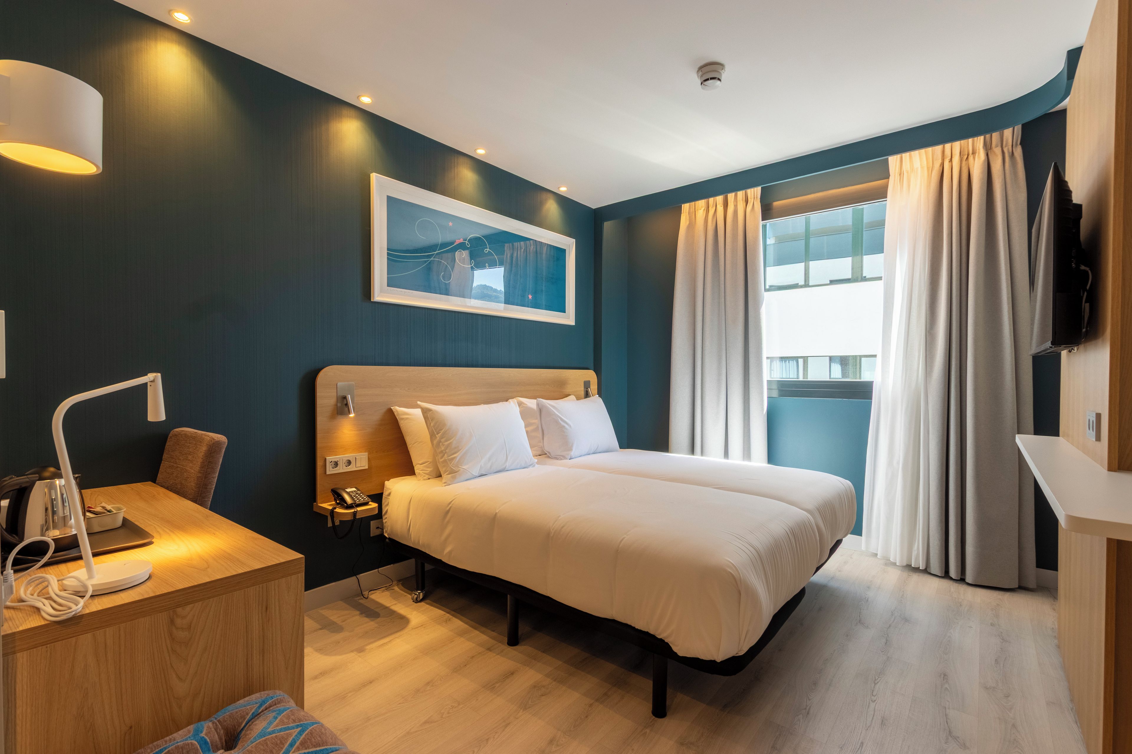 A hotel room with a bed, bedside table, lamp, and a large window with curtains.
