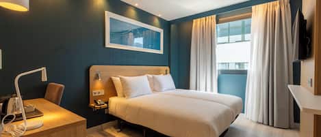 Habitación de hotel con cama, mesita de noche, lámpara y un amplio ventanal con cortinas.