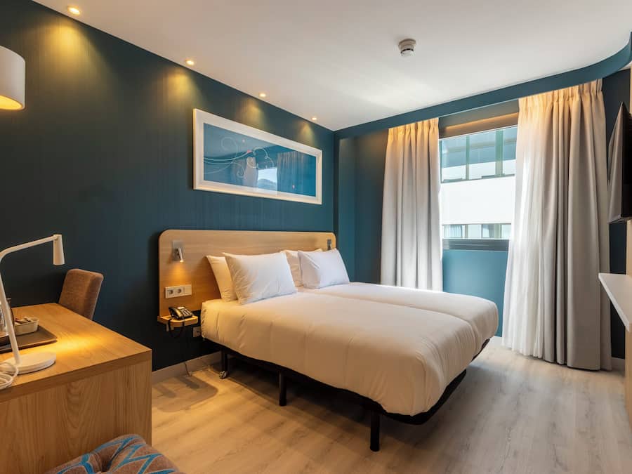 Habitación de hotel con cama, mesita de noche, lámpara y un amplio ventanal con cortinas.
