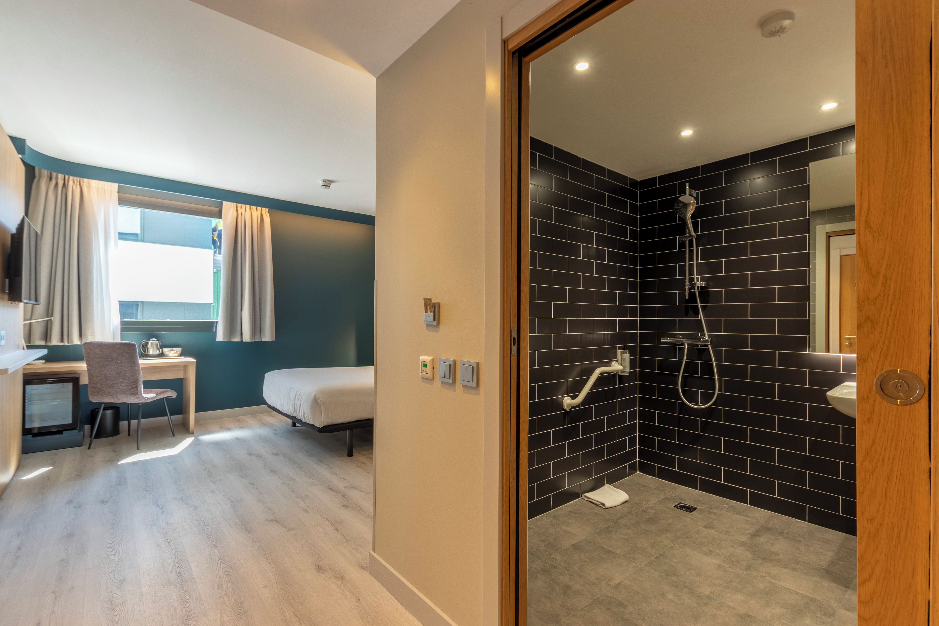 A modern hotel room with a desk, chair, bed, and a shower area with black tiles.