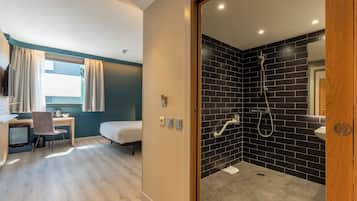 A modern hotel room with a desk, chair, bed, and a shower area with black tiles.
