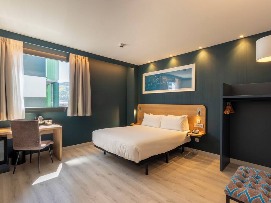 Una habitación de hotel moderna con cama, escritorio, silla y ventana con cortinas.