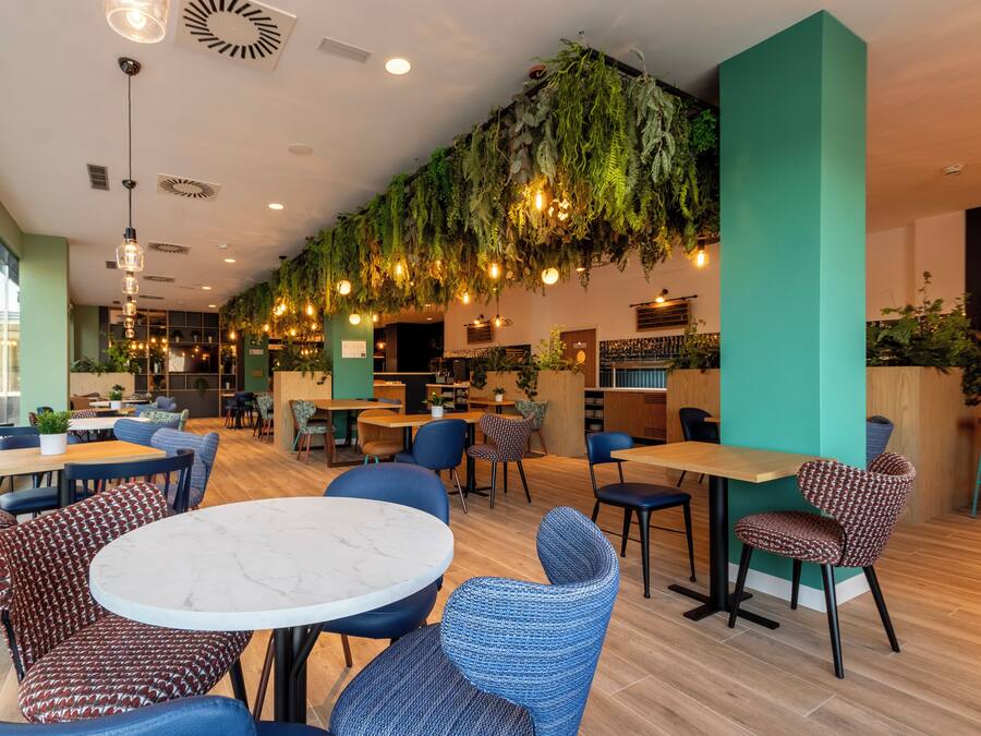 A modern cafe with round tables, patterned chairs, hanging plants, and a green accent wall.