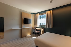 Ein Hotelzimmer mit Bett, Schreibtisch mit Stuhl, Fernseher und einem Fenster mit Vorhängen.