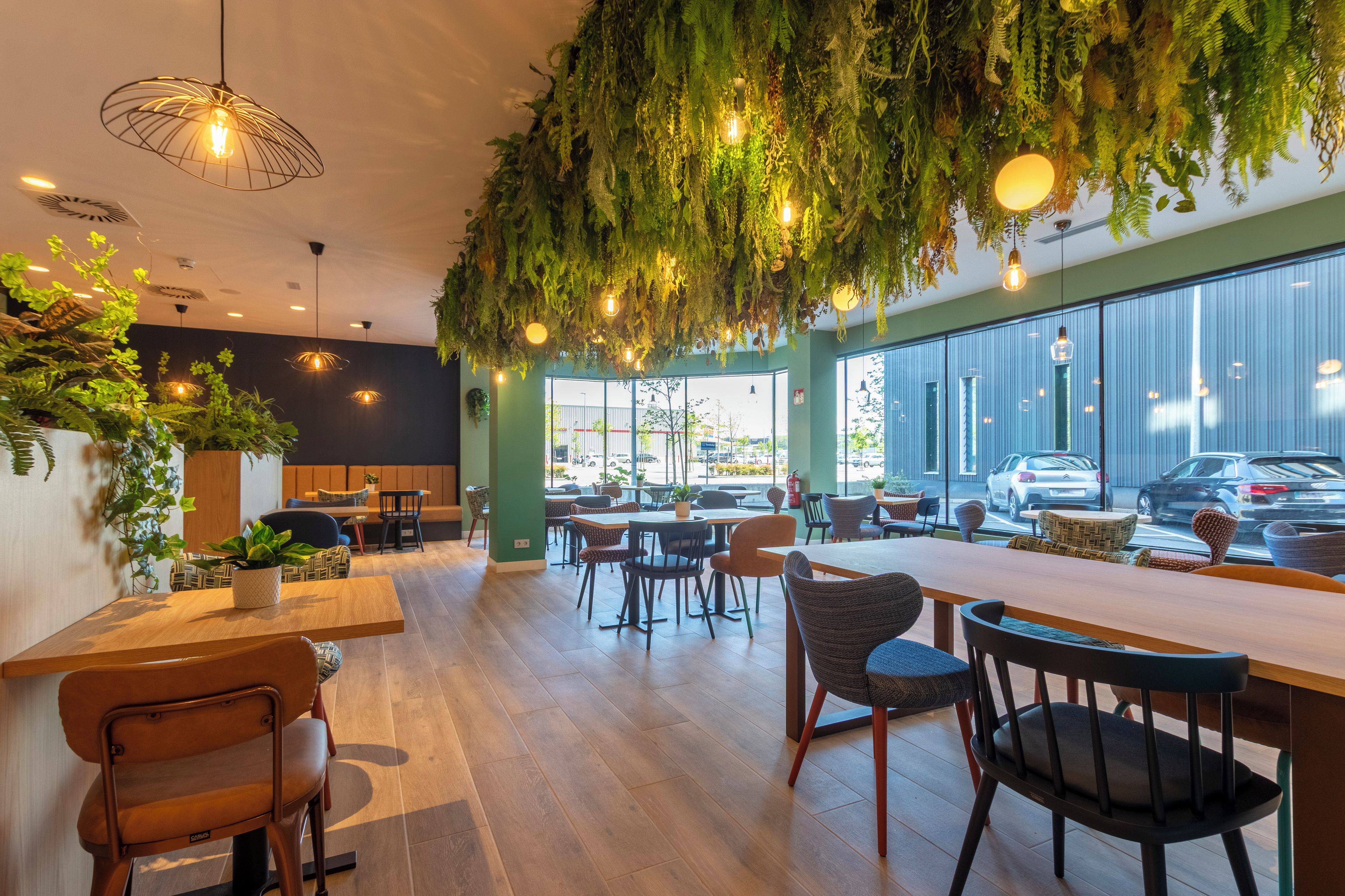 A modern interior with wooden tables, chairs, hanging plants, and large windows.