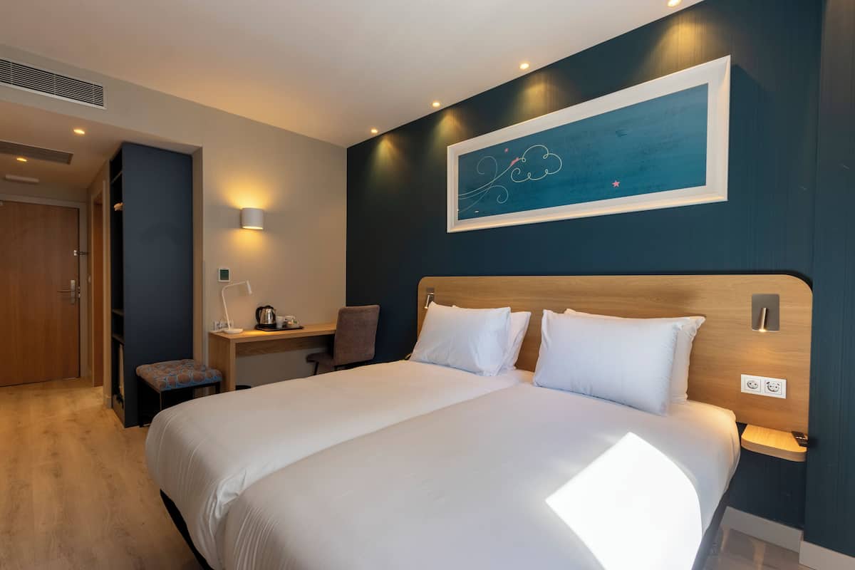Een hotelkamer met een groot bed, een bureau met stoel, een kunstwerk aan de muur en een klein bankje.