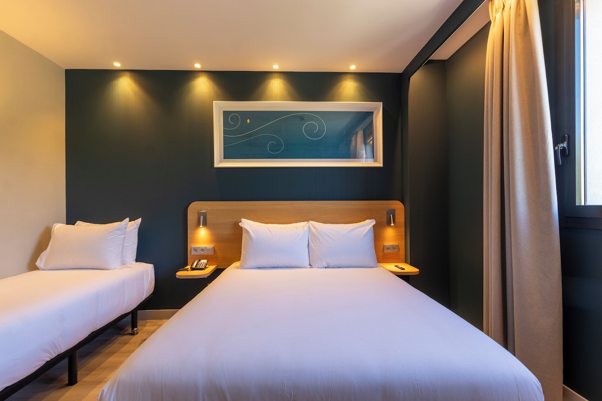 Een hotelkamer met twee bedden, een hoofdeinde, bedlampen, een kunstwerk aan de muur en een raam met gordijnen.