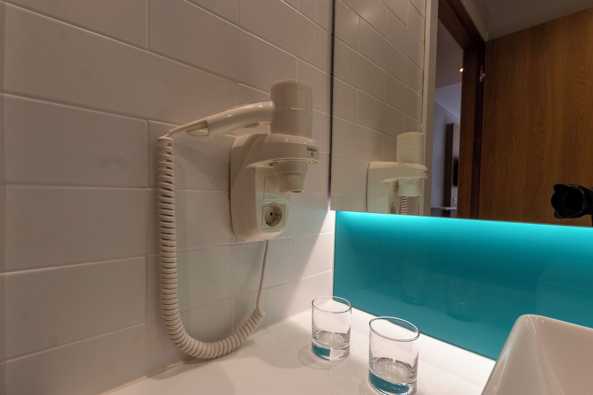 Casa de banho com secador de cabelo fixado na parede, espelho e dois copos de água no lavatório.