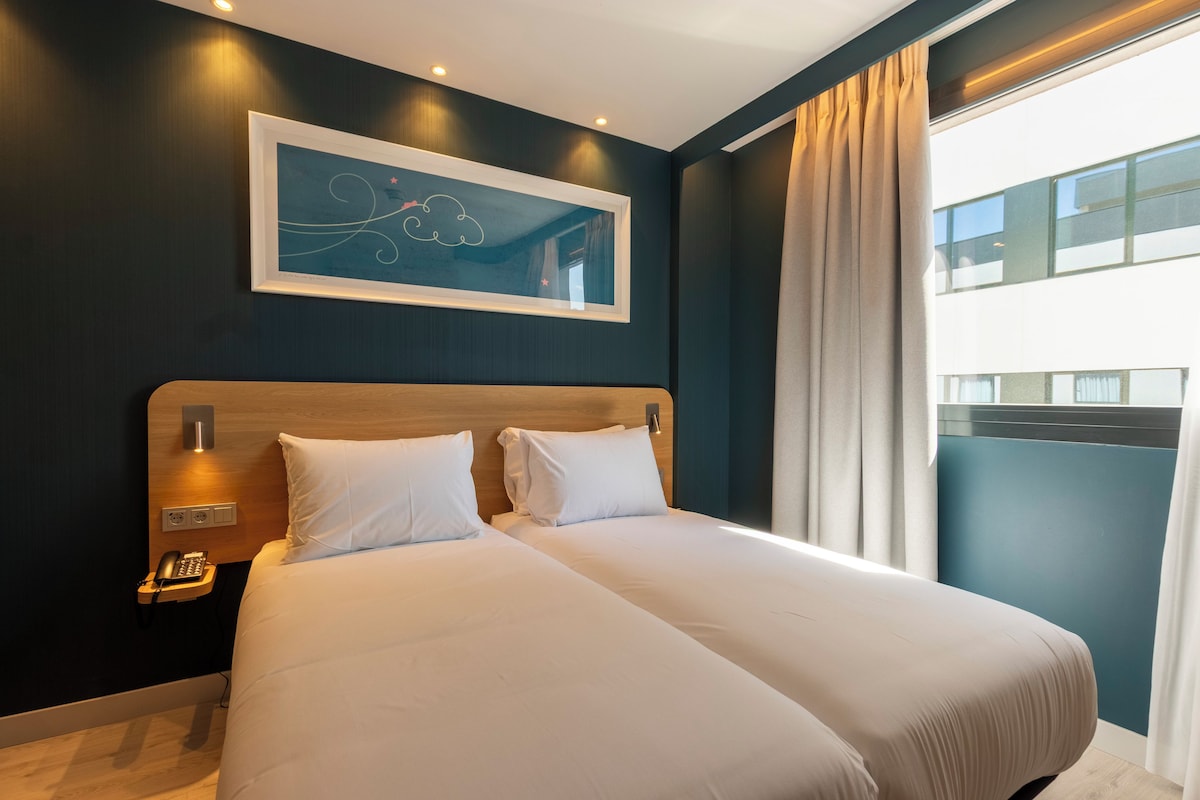 Een hotelkamer met een groot bed, een bedtelefoon, een ingelijst kunstwerk aan de muur en uitzicht op een gebouw door het raam.