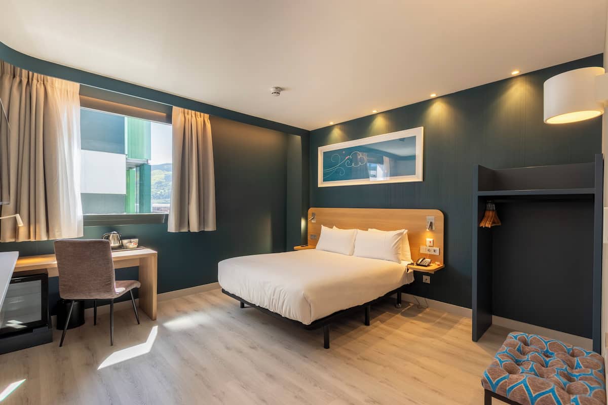 Een moderne hotelkamer met een bed, bureau, stoel en een raam met gordijnen.