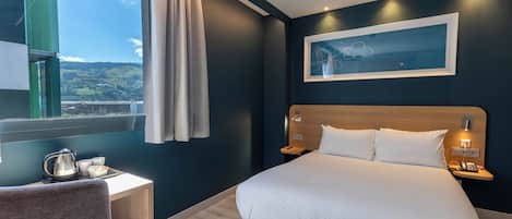 Habitación de hotel moderna con cama, mesitas de noche, escritorio con hervidor y vistas al exterior.