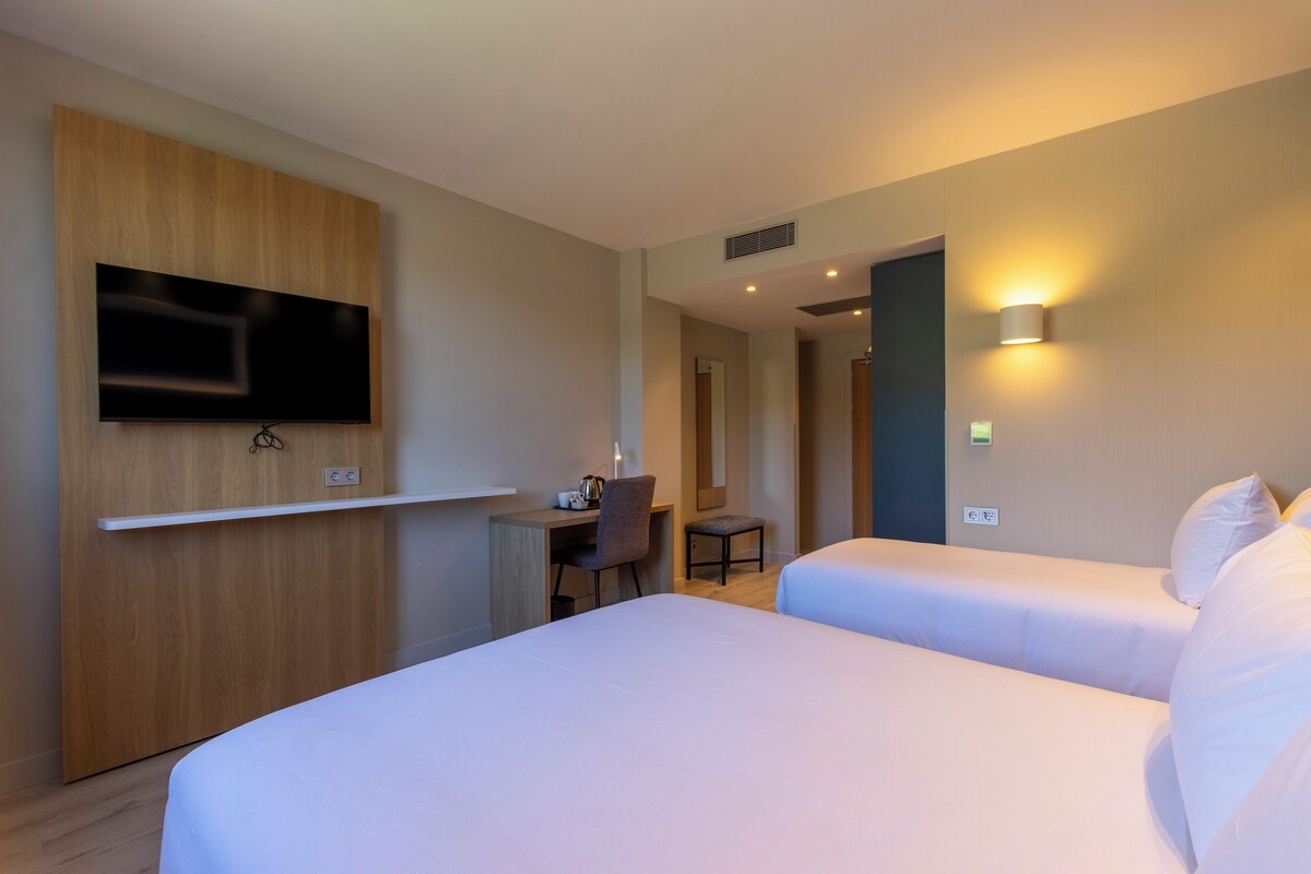 Quarto de hotel com cama, escrivaninha, cadeira, televisão na parede e luminária de parede.