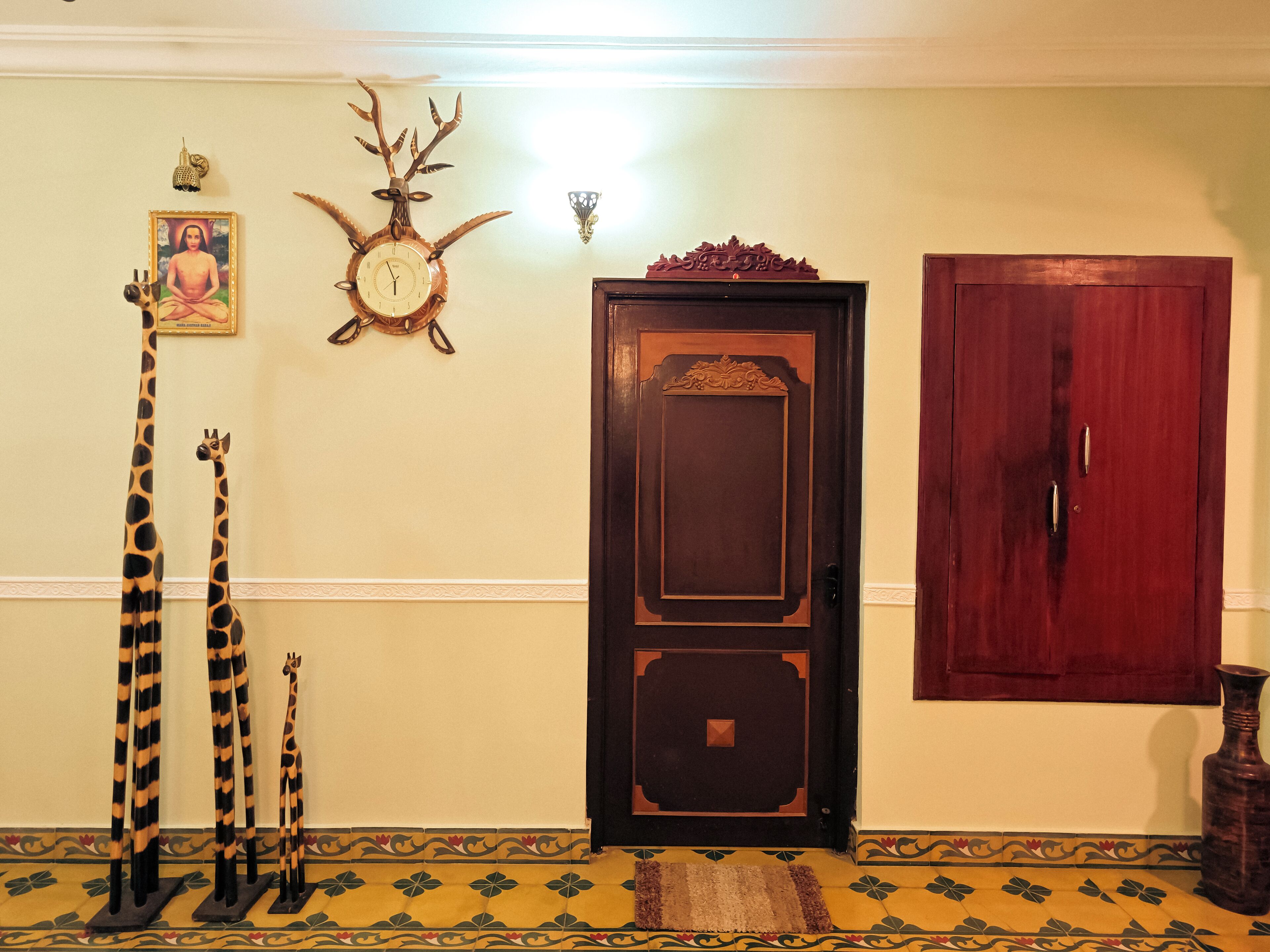 A room with a wooden door, a wall-mounted clock, a framed picture, and two giraffe-shaped sculptures.