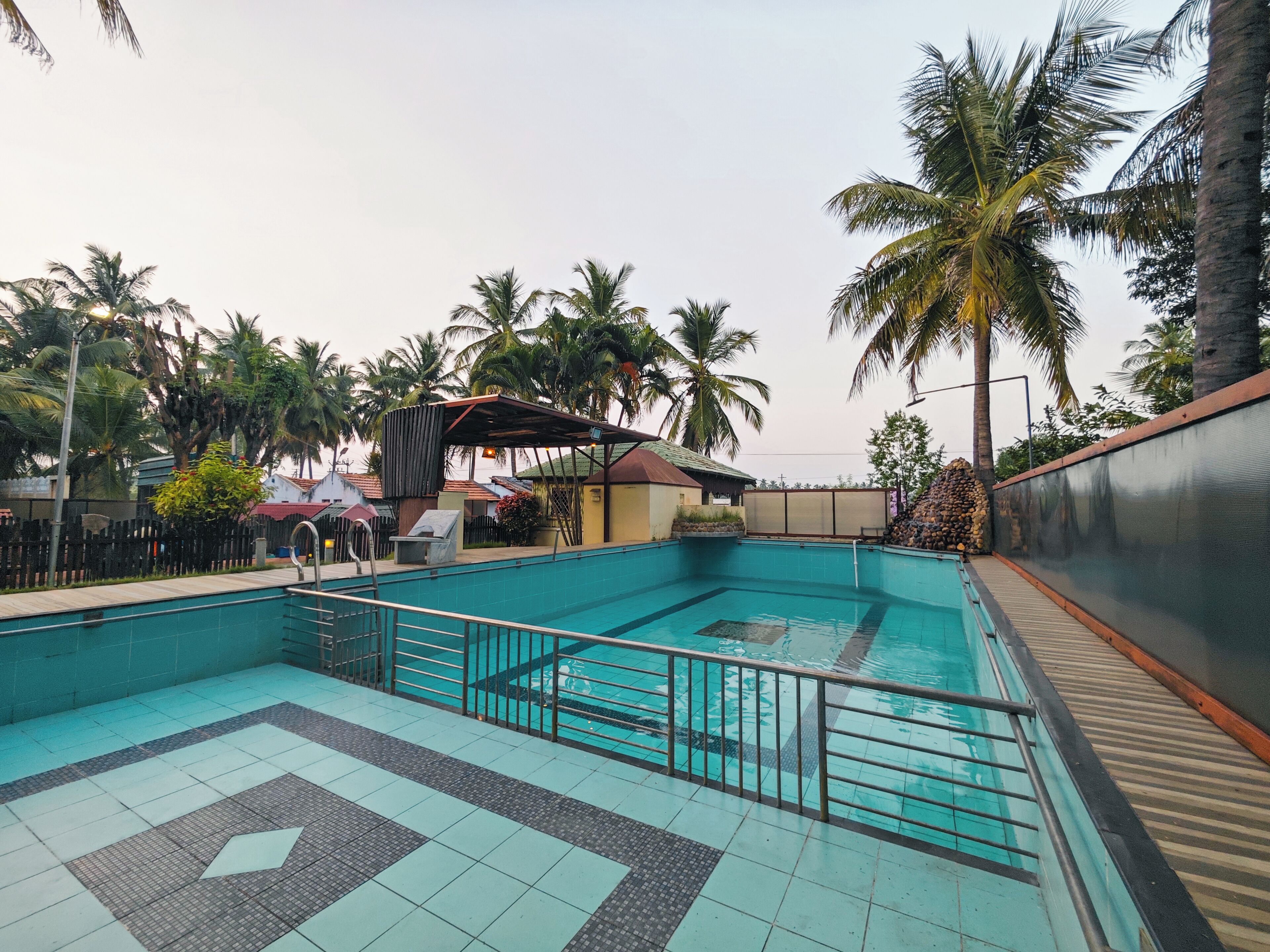 A swimming pool with tiled flooring, surrounded by palm trees and a fence.