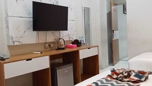 A hotel room with a wooden desk, a television mounted above it, a small refrigerator, a bed with a patterned blanket, and a mirror. - Edu Hotel Y-skanam (Surabaya)