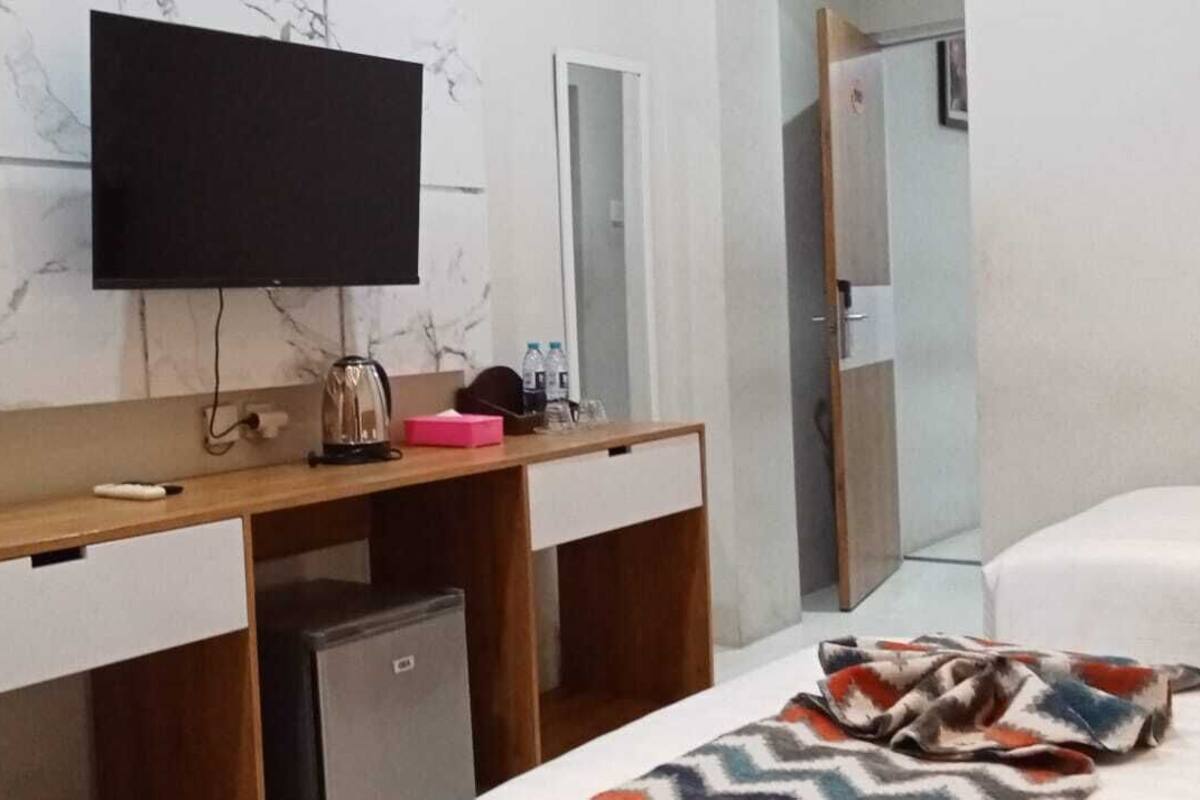 Habitación de hotel con un escritorio de madera, un televisor montado encima, un refrigerador pequeño, una cama con un edredón estampado y un espejo.